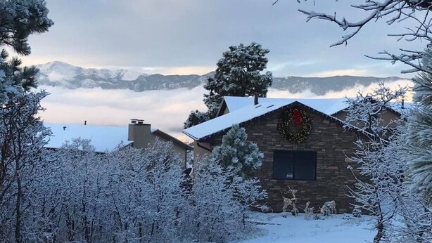 Una cabaña rústica de fachada de piedra, rodeada de árboles cubiertos de nieve y una vista despejada de montañas distantes.
