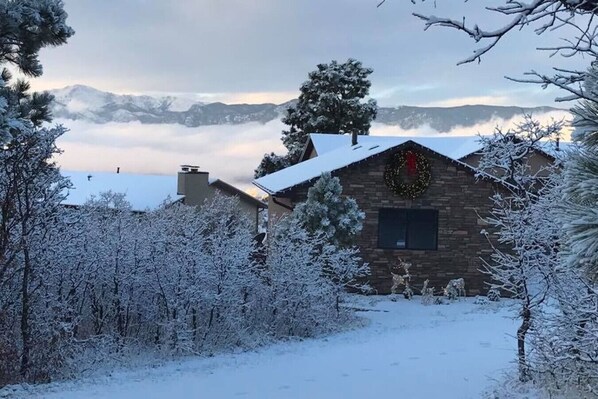 Una cabaña rústica de fachada de piedra, rodeada de árboles cubiertos de nieve y una vista despejada de montañas distantes.