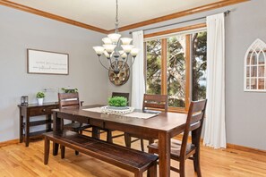 A dining room with a wooden table, chairs, and a chandelier. There is a window with curtains, a framed quote on the wall, and a small plant on the table.