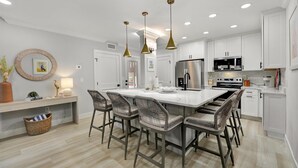 Une cuisine moderne avec des meubles blancs, un îlot central doté de tabourets de bar, un éclairage suspendu et des appareils en acier inoxydable.