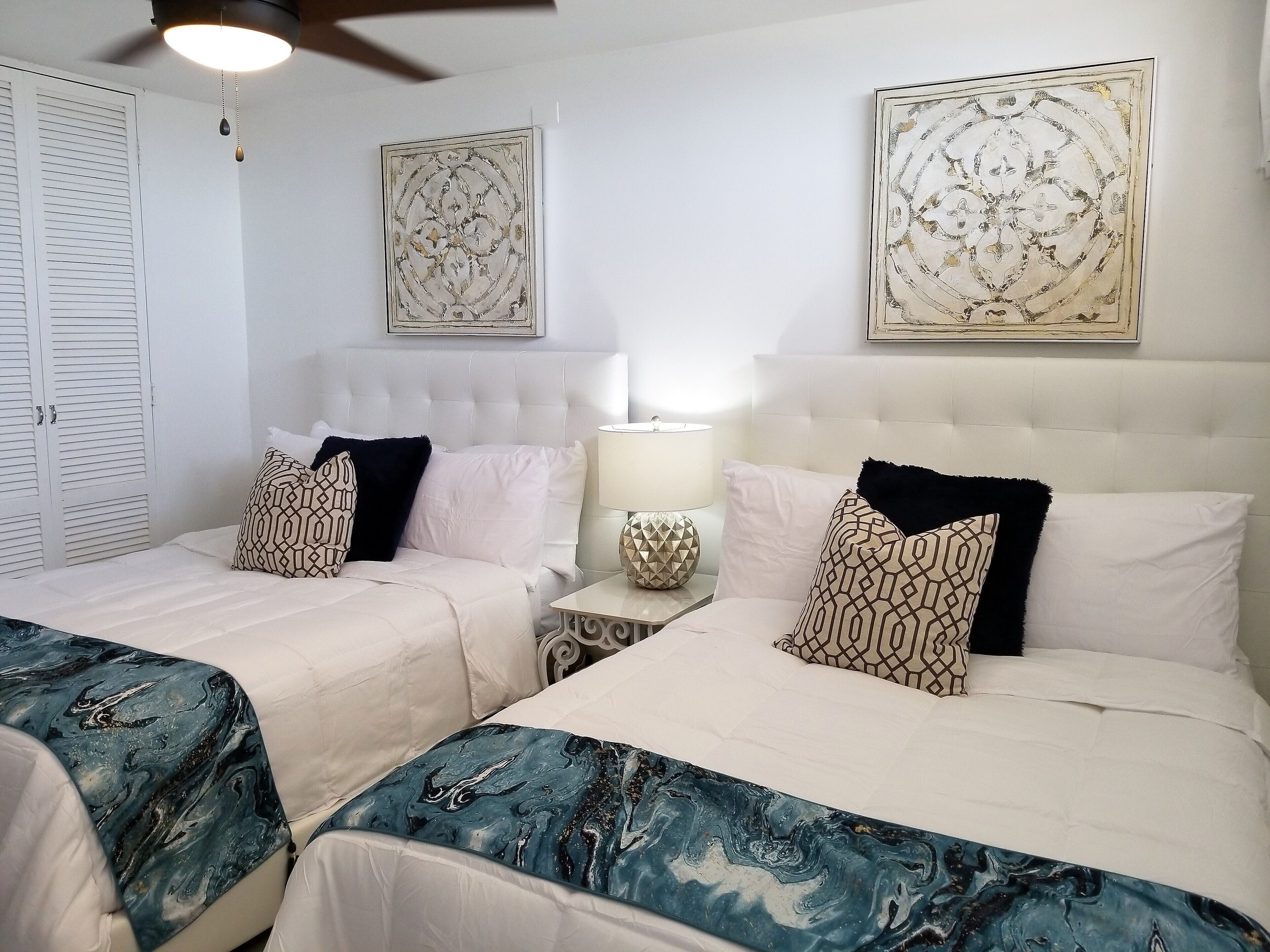 A hotel room with two beds, a ceiling fan, a bedside table with a lamp, and two wall-mounted artworks.