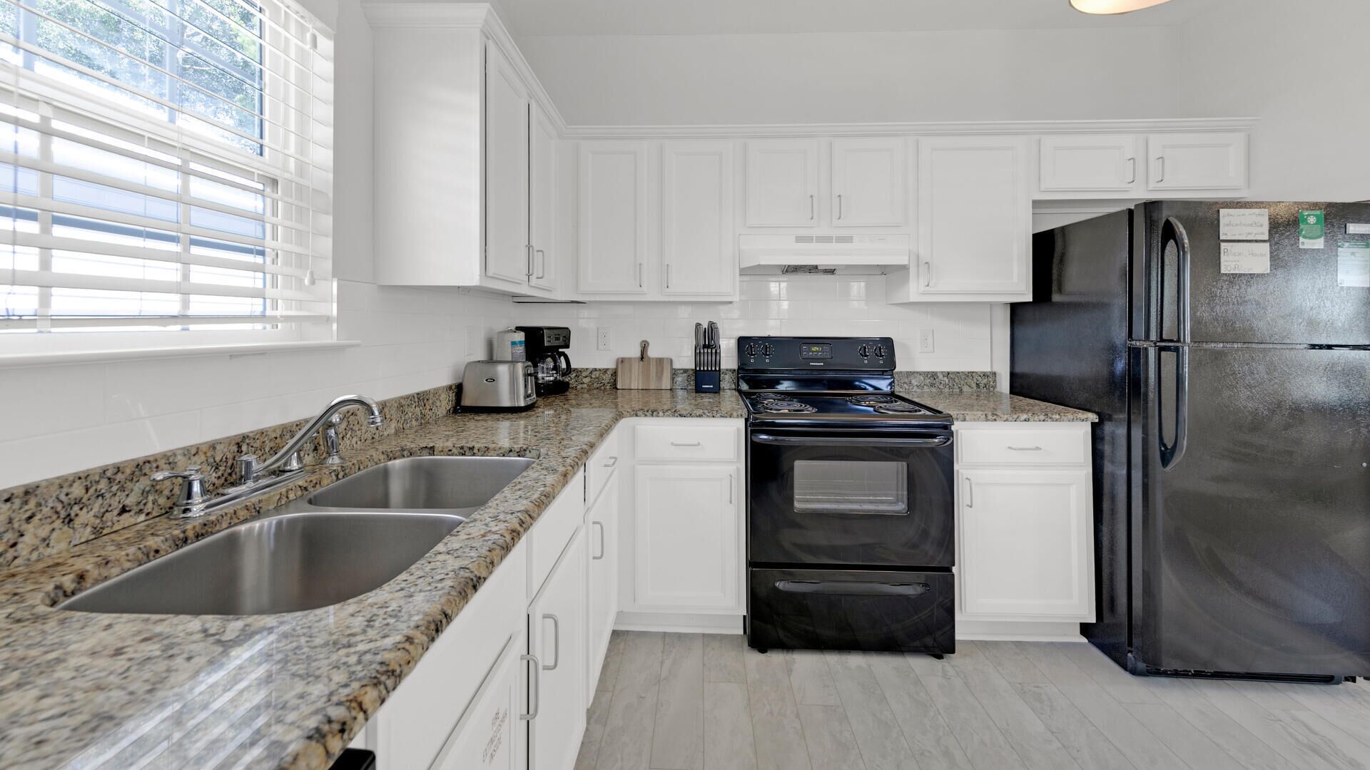 A modern kitchen with white cabinets, granite countertops, stainless steel appliances, and a window with blinds.