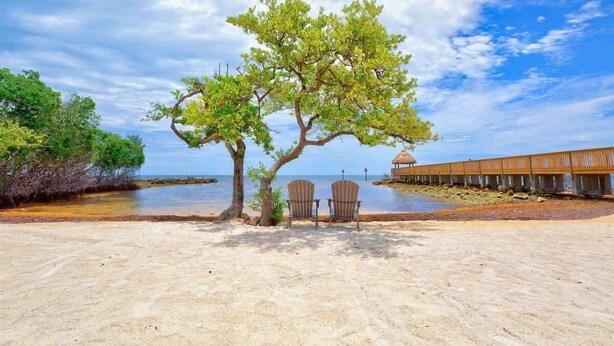 A beach with two chairs under a tree, a wooden structure, and a pier in the background. - Charming & Tranquil Condo at Ocean Pointe-Sleeps 6 (Tavernier)