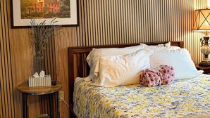 Une chambre d’hôtel avec un mur en lambris de bois, un lit avec une literie blanche, une couvre-lit à motifs floraux, une table de chevet avec une lampe et un tableau encadré au-dessus du lit.