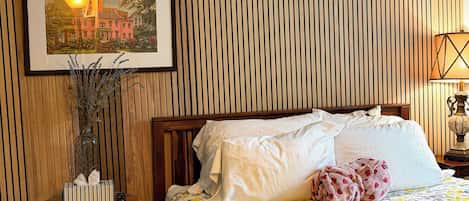 A hotel room with a wooden slat wall, a bed with white bedding, a floral-patterned bedspread, a bedside table with a lamp, and a framed picture above the bed.