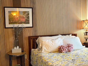 Habitación de hotel con pared de listones de madera, cama con ropa blanca, colcha de estampado floral, mesita de noche con lámpara y un cuadro enmarcado sobre la cama.