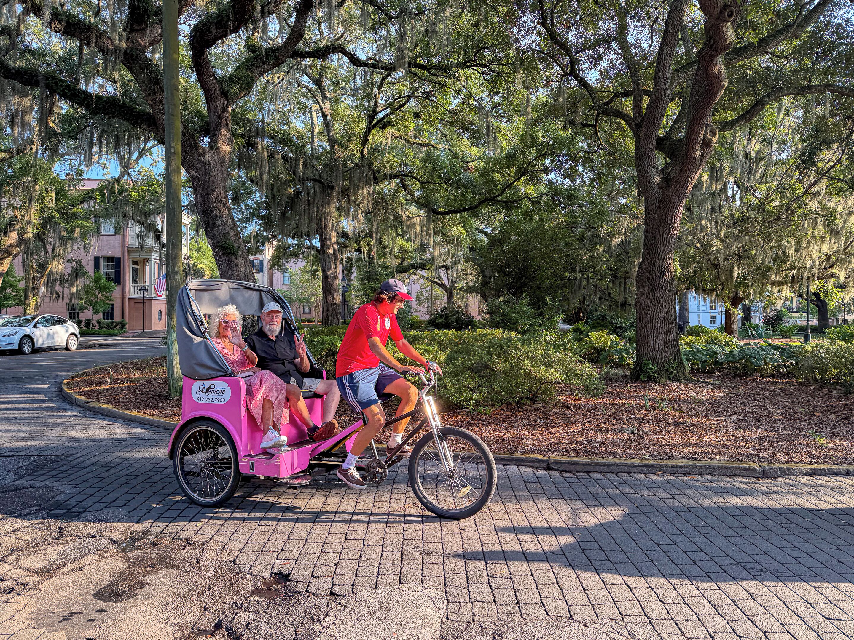 A pink trolley with passengers, parked on a paved path surrounded by trees and greenery.