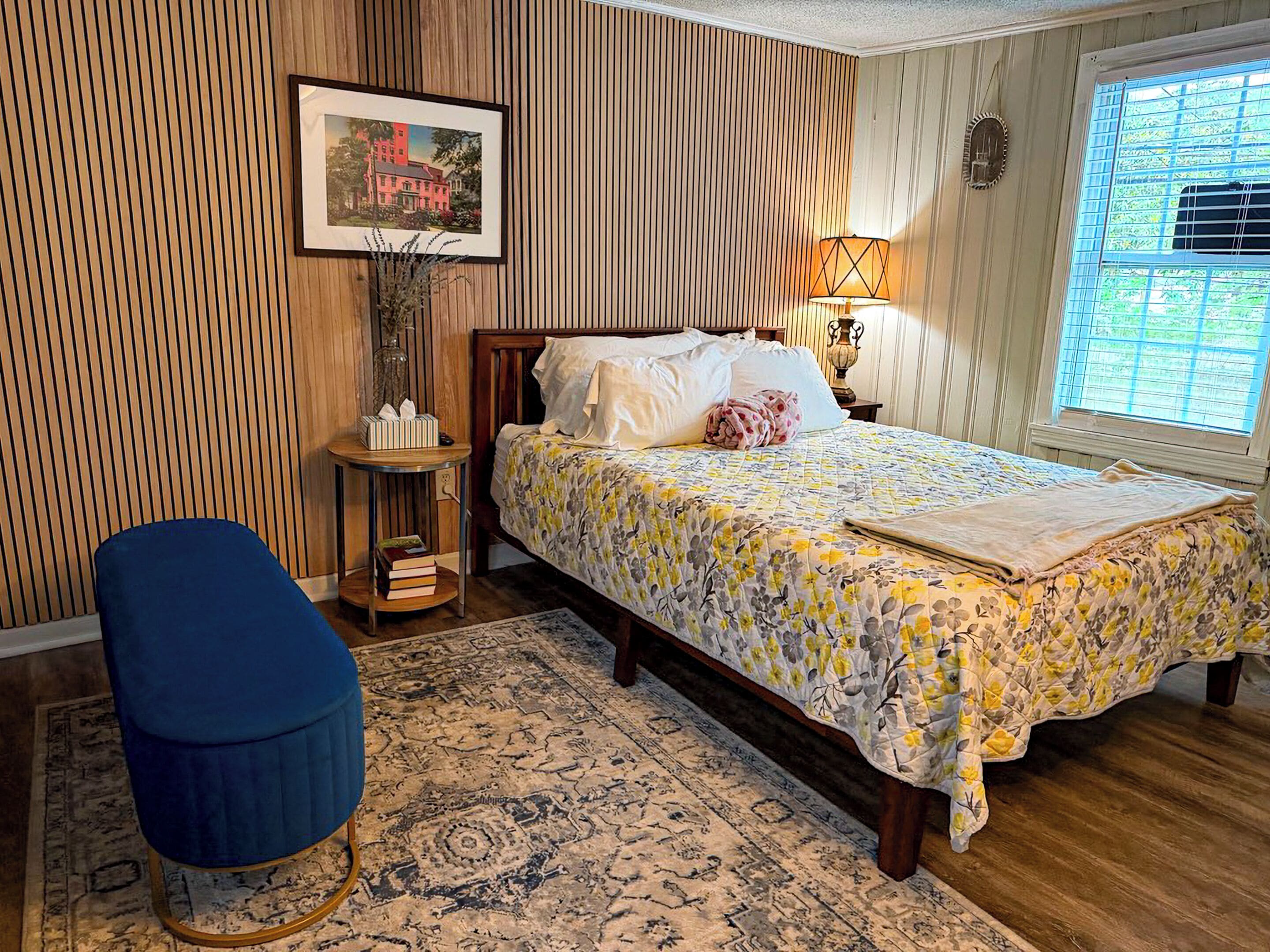 Un dormitorio con cama, mesita de noche, lámpara, silla, alfombra y ventana con persianas.