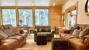 A cozy cabin interior with leather sofas, a wooden dining table, and large windows offering a forest view.