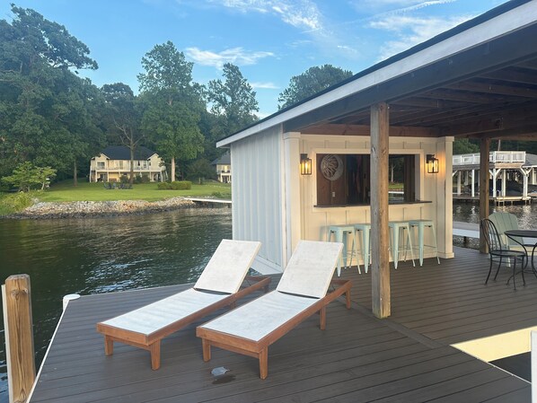 A wooden deck with lounge chairs and a covered seating area overlooking a body of water, with a dock extending into the water.