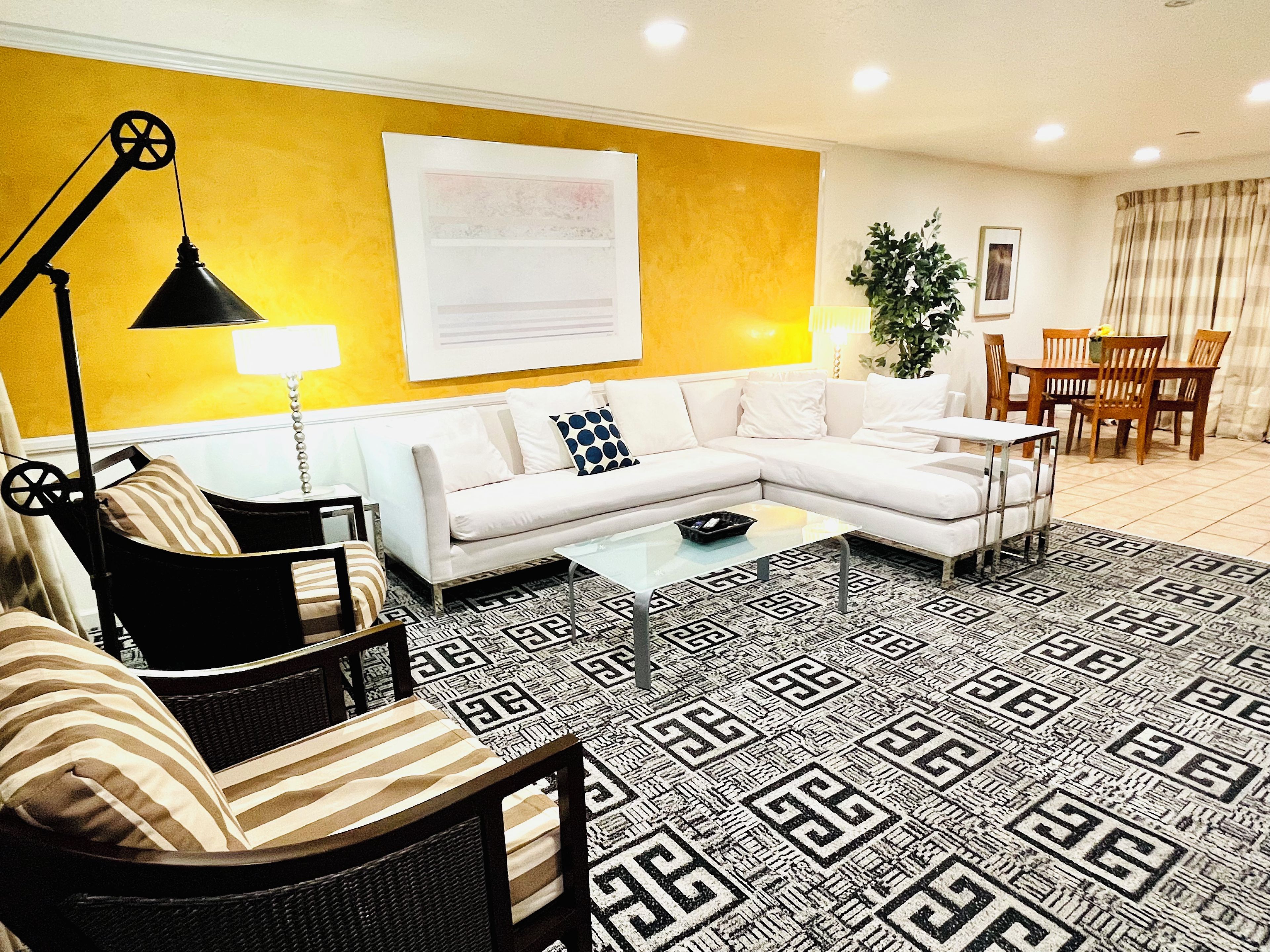 A living room with a white sofa, a black floor lamp, a patterned rug, and a dining area with wooden chairs and a table.
