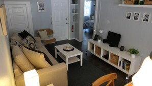 A living room with a sofa, armchair, coffee table, and TV stand. There is a door leading to another room and a shelf with decorative items. - Dream holiday at a great price – now with massive discounts! (Fort Myers)