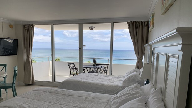 Et hotelværelse med to senge, et fjernsyn, en balkon med bord og stole og udsigt til havet.