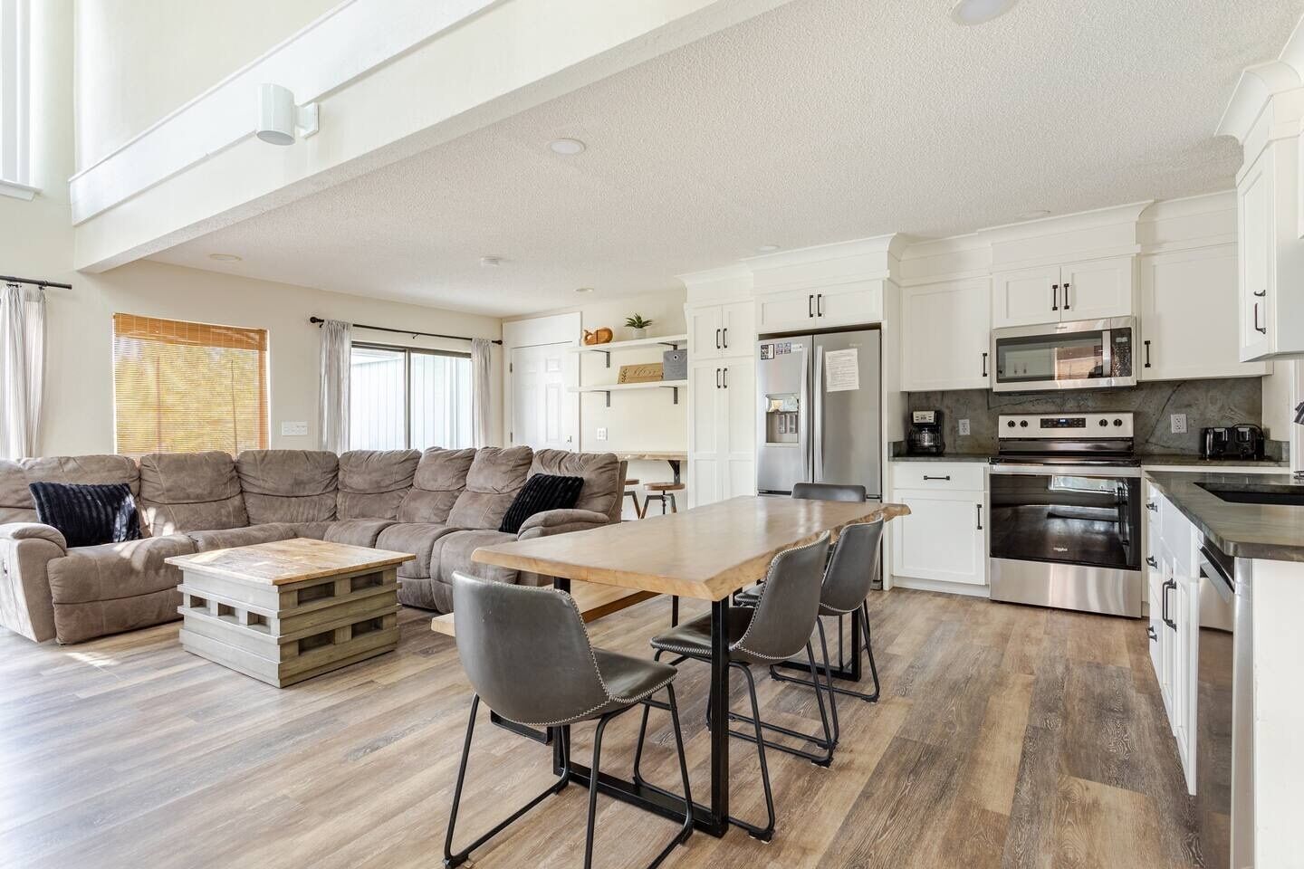A spacious living area with a sofa, a wooden coffee table, and a dining table with chairs. There is a kitchen with modern appliances and white cabinetry.