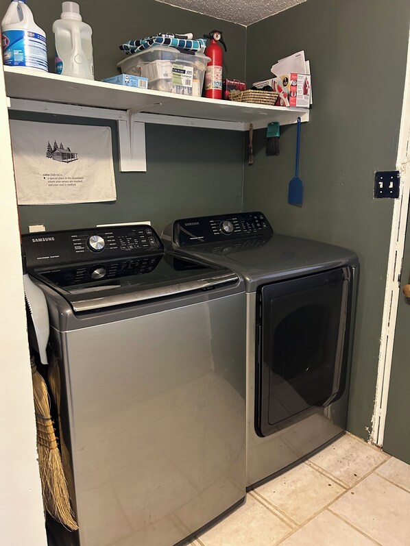 A laundry area with two Samsung washing machines, a broom, a fire extinguisher, and cleaning supplies on a shelf. - Family-Friendly, Cozy, Spacious Cabin Outside Of Hot Springs, NC - Just Magical (Hot Springs)