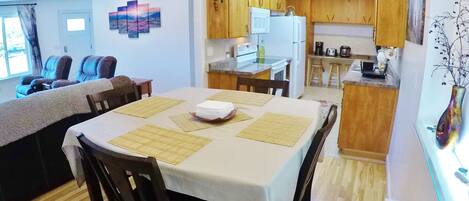 Un comedor con una mesa dispuesta para cuatro personas, sillas de madera y una cocina al fondo con armarios y electrodomésticos de madera.