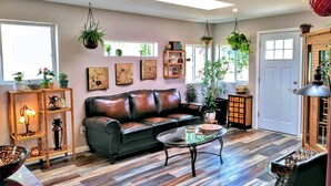A living room with a leather sofa, a glass coffee table, and various plants. There are framed artworks on the wall and a wooden bookshelf. - Reconnect ~ Relax ~  Rejuvenate ~ Explore ~  Larch Avenue Getaway (Port Angeles)