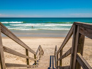 Une promenade en bois mène à une plage de sable, avec l’océan en arrière-plan.