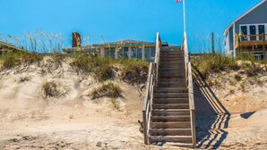 A wooden staircase leading to a building with a flag on top, surrounded by sand and sparse vegetation.