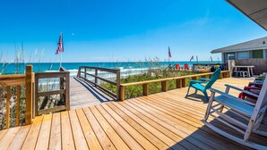 A wooden deck with a view of the ocean, featuring a beach house, beach chairs, and American flags.