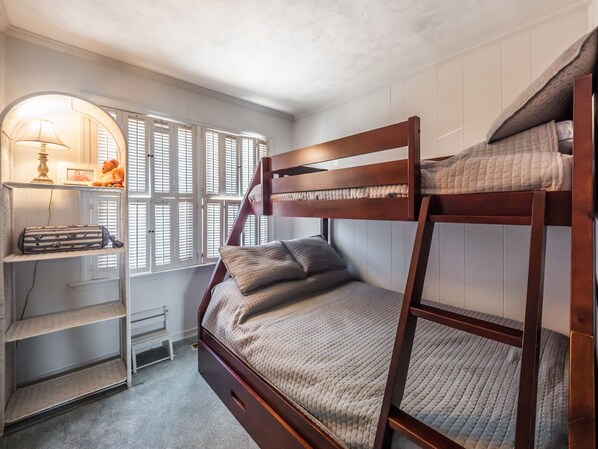 A bunk bed room with a desk and shelves, a window with blinds, and a lamp.