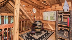 Interior de una cabaña de madera con una escalera, un barril grande y una ventana.