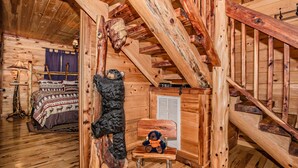 Interior de una cabaña de madera con una escalera de madera, una cama con ropa de cama estampada y un osito de peluche en una silla.