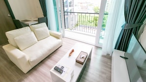 A modern living room with a white sofa, a coffee table with a remote and a book, and a balcony with a view of buildings and greenery. - Locals ChiangMai Central Airport Plaza (Chiang Mai)