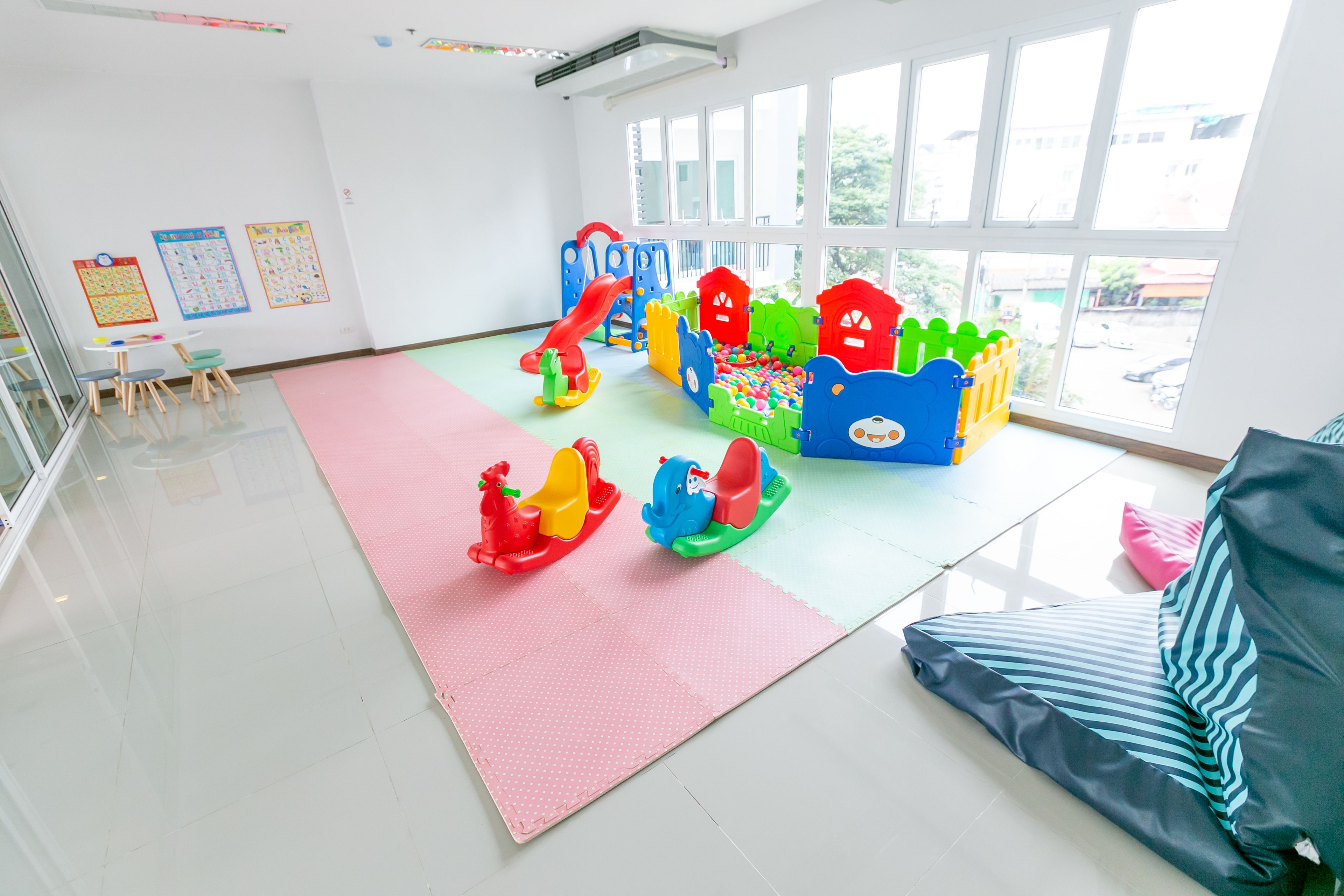 Una habitación luminosa con ventanas amplias, una zona de juegos con juguetes coloridos y un tapete rosa en el suelo.