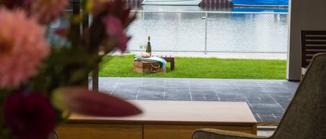 A view from a room with a table and a wicker basket containing a bottle and glasses, overlooking a grassy area with a fence and boats on the water.