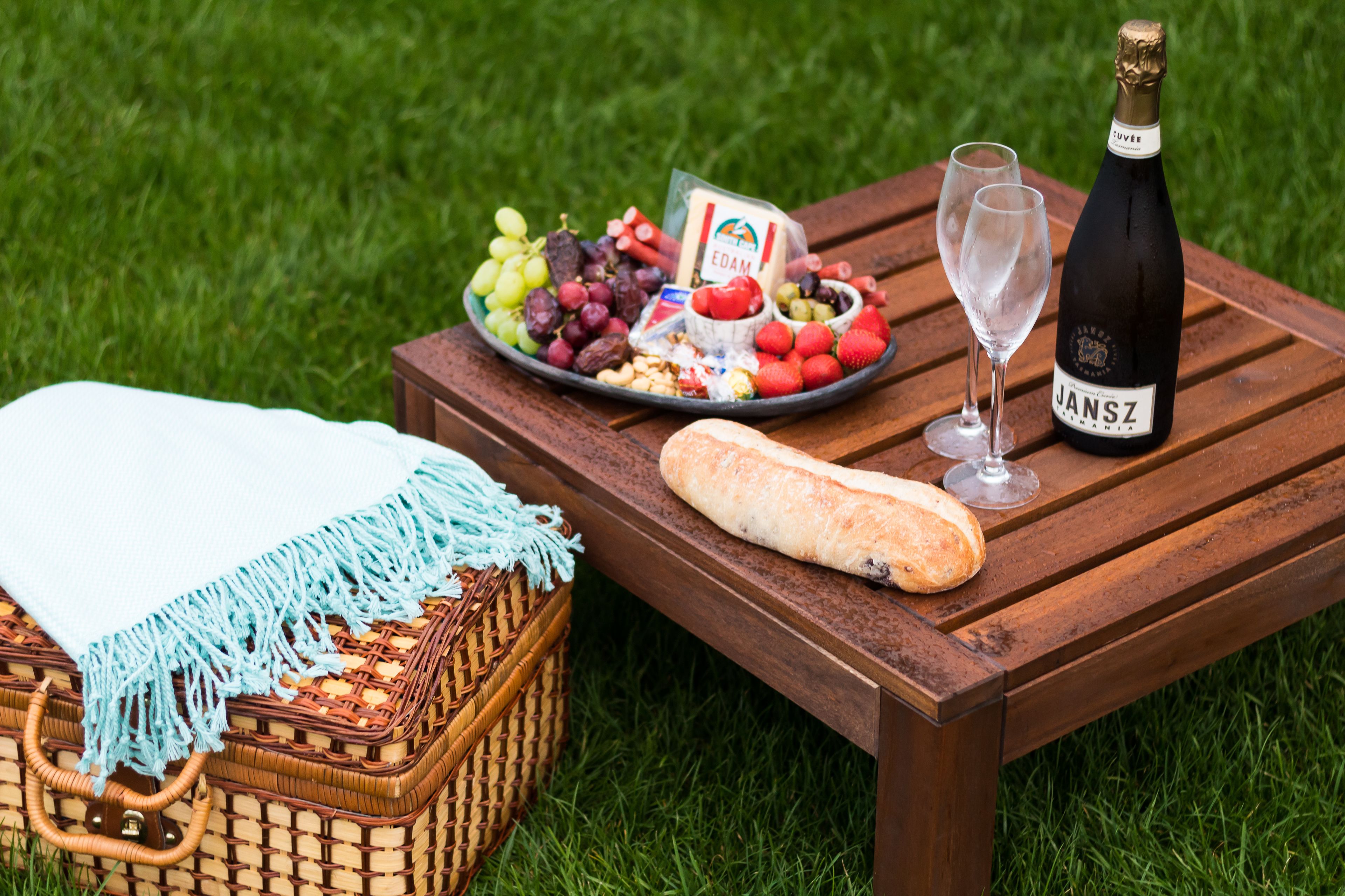 Un montaje para picnic sobre una mesa de madera con una botella de champán Jansz, una cesta y un plato de frutas y queso.