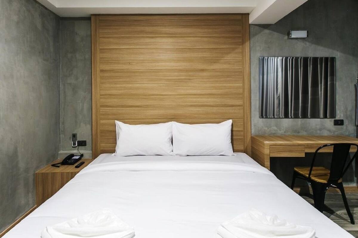A modern hotel room with a wooden headboard, a bed with white linens, a bedside table with a phone, and a desk with a chair.