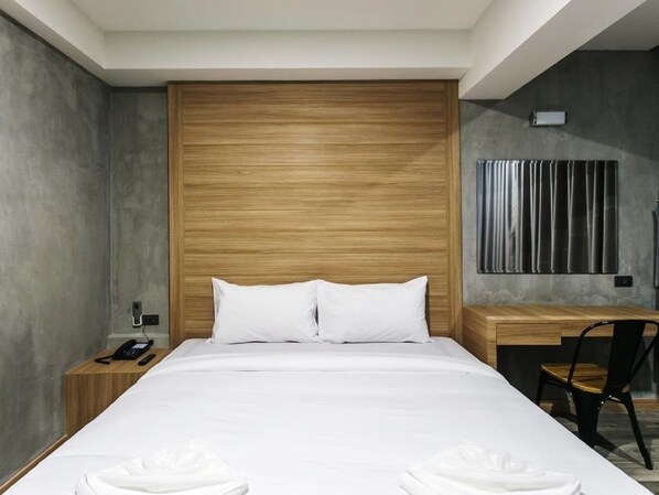 A modern hotel room with a wooden headboard, a bed with white linens, a bedside table with a phone, and a desk with a chair. - Happy Bou Jomtien Guesthouse (Pattaya)
