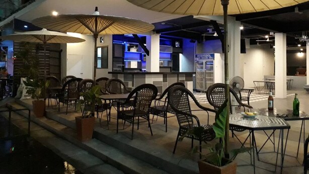 An outdoor dining area with tables and chairs, umbrellas, and a view of the building's interior. - Happy Bou Jomtien Guesthouse (Pattaya)
