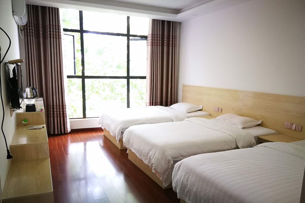 A hotel room with two beds, a wooden nightstand, a window with curtains, and a view of greenery outside.