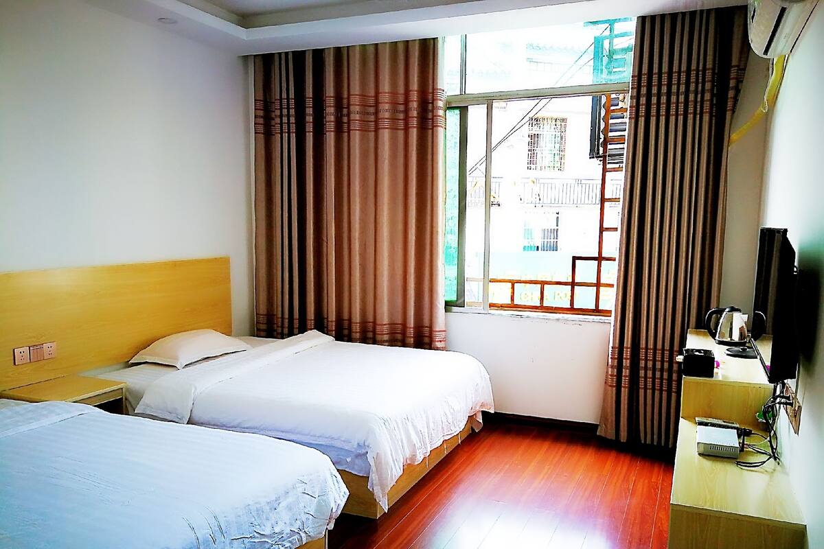 A hotel room with two beds, a wooden floor, a window with curtains, a desk with a computer, and a TV.