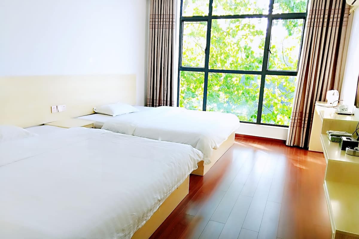 A hotel room with two beds, a wooden floor, a window with curtains, and a desk with a lamp.