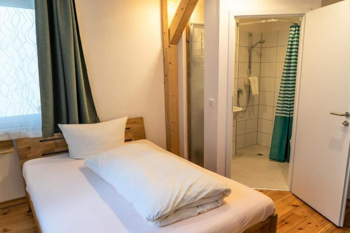 A bedroom with a wooden bed, white bedding, a window with a curtain, and a shower area visible through an open door.
