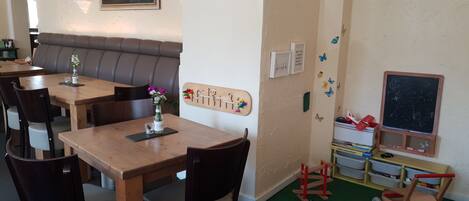 A dining area with wooden tables and chairs, a cushioned bench, a chalkboard, and a toy cart.