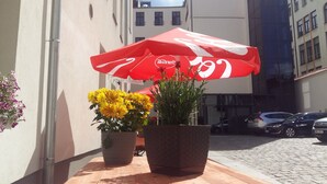 A red umbrella with a Coca-Cola logo, two potted plants with yellow flowers, and a cobblestone street with parked cars. - Kamienica Pod Aniolami (Wroclaw)