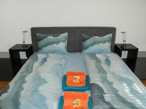 Ein Bett mit grauem Kopfteil, zwei passende Kissen und eine blaue und weiße Bettdecke. Auf beiden Seiten des Bettes befinden sich jeweils ein schwarzer Nachttisch mit einer Lampe.
