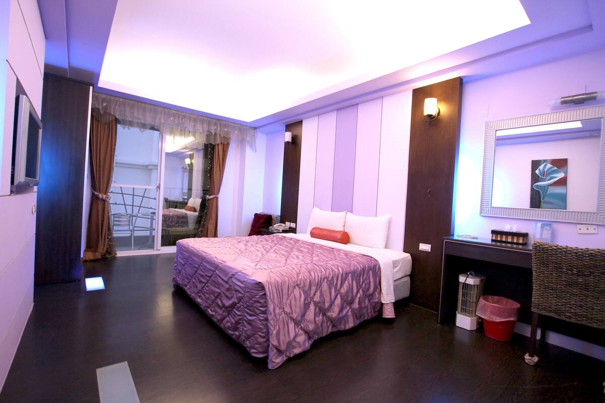 A modern hotel room with a large bed, a small desk, a mirror, and a sliding door leading to a balcony.