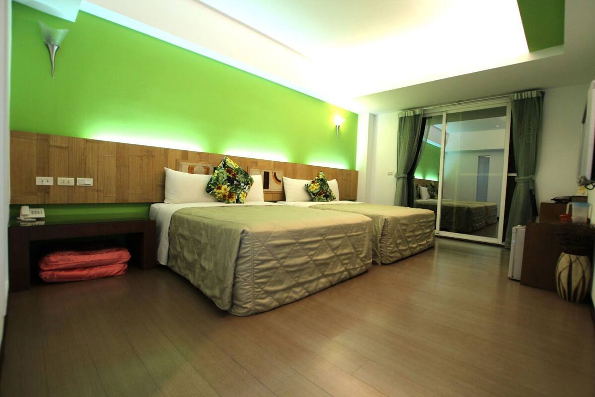 A hotel room with two beds, a wooden headboard, a green accent wall, a sliding glass door, and a desk with a lamp.