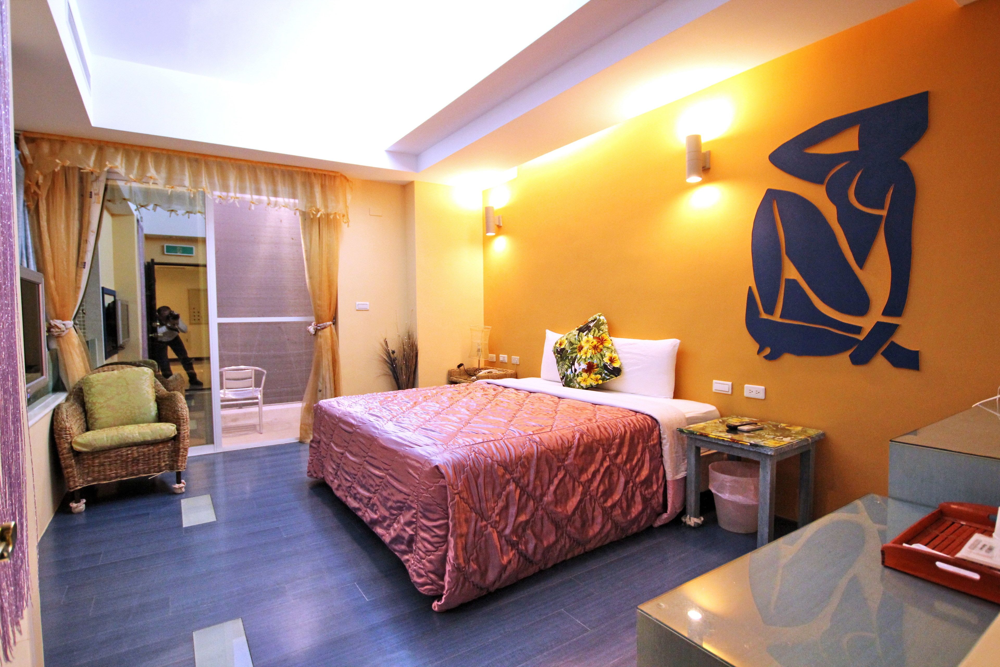 A hotel room with a large bed, a small table, a chair, and a wall-mounted light fixture.