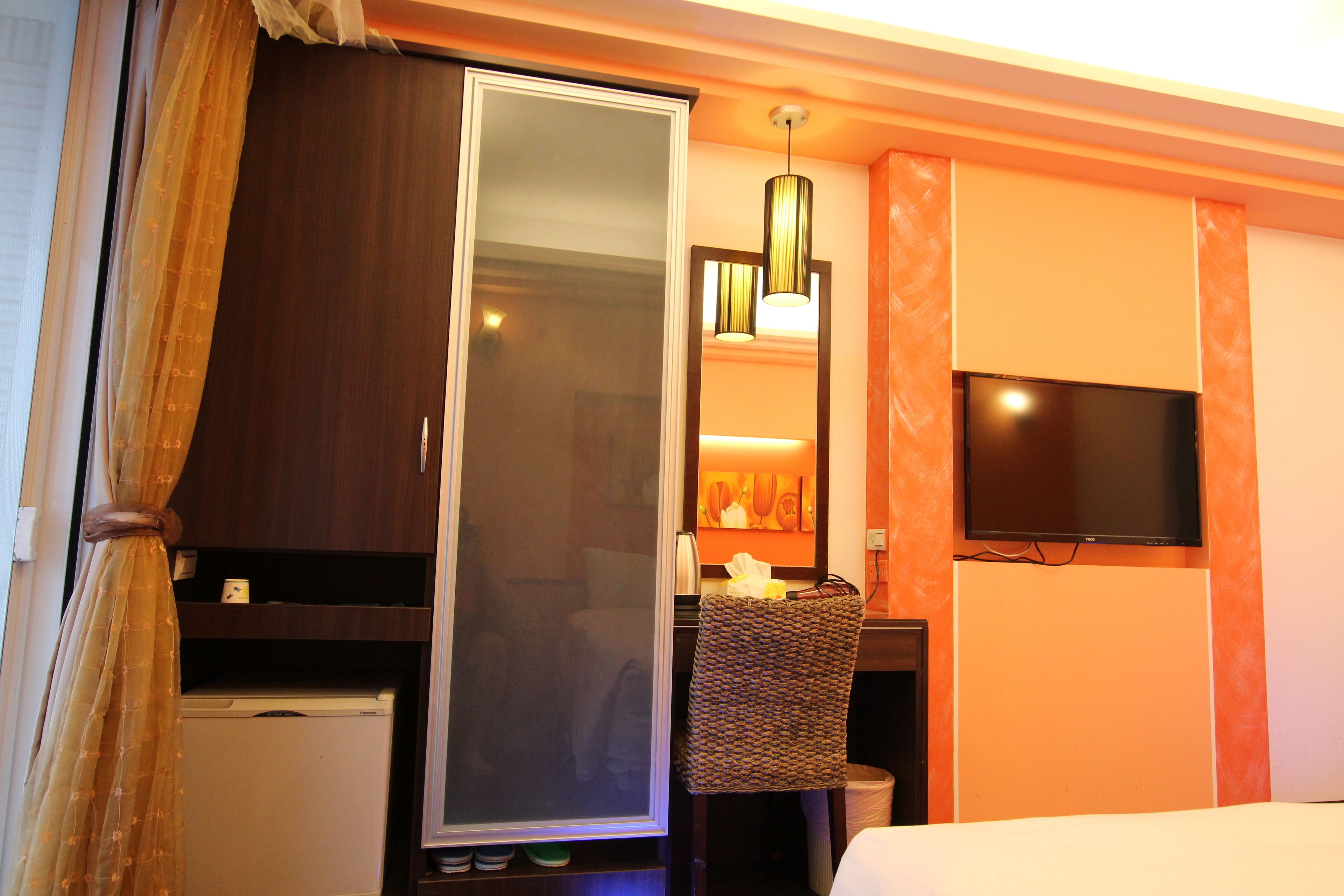 A hotel room with a wardrobe, a television mounted on the wall, a small table with a lamp, and a bed.