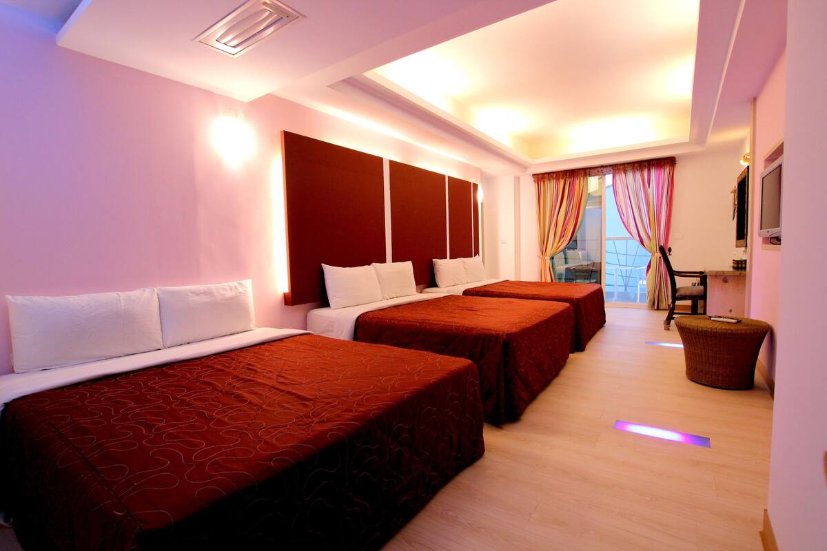 A hotel room with three beds, a flat-screen TV, and a large window with curtains.