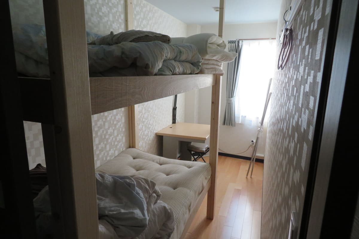 A bunk bed room with a desk and chair, a window with curtains, and a door with a handle.
