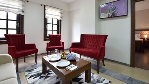 A room with a dining table set for two, two red armchairs, a white sofa, and a television mounted on the wall. - Zagnospasa Konaklari (Trabzon)