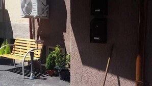 Un edifici amb dues unitats de aire condicionat, una banca de fusta, plantes en testos i una escombra adossada a la paret.