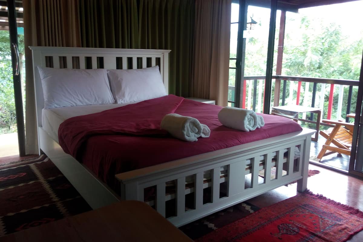 A bed with white headboard and pillows, a red bedspread, and two rolled towels.